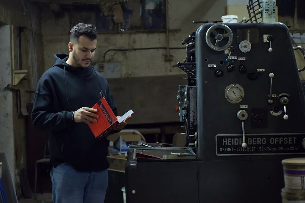 A man in a black hoodie reads by an industrial printing machine in Benghazi, Libya.